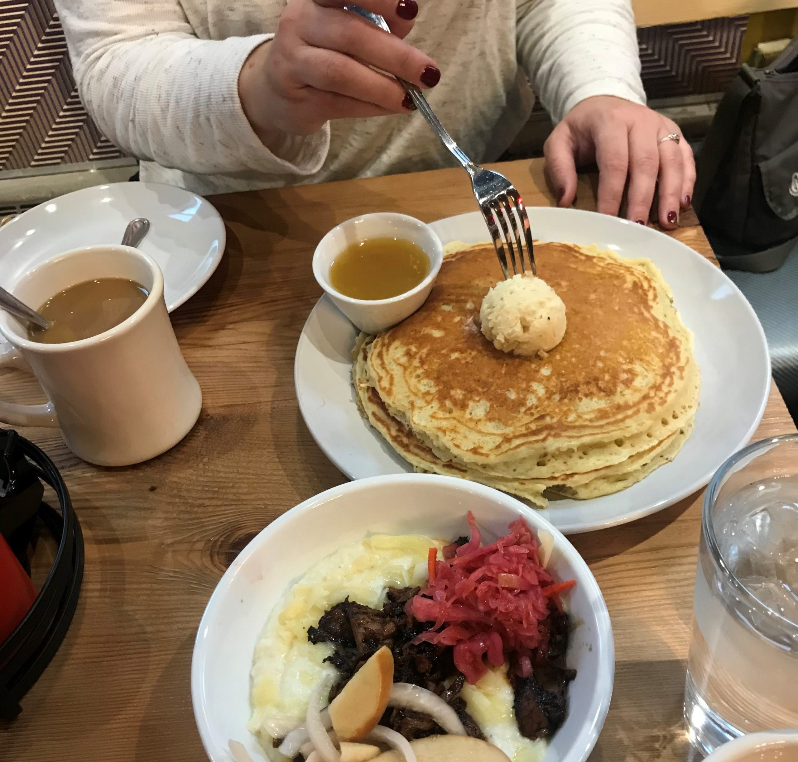 A hand cutting into pancakes with a fork, surrounded by other breakfast dishes.