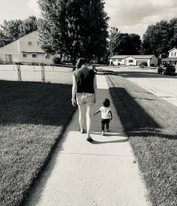 Black and white photo of mom walking down sidewalk with toddler. Photo is taken from behind. 